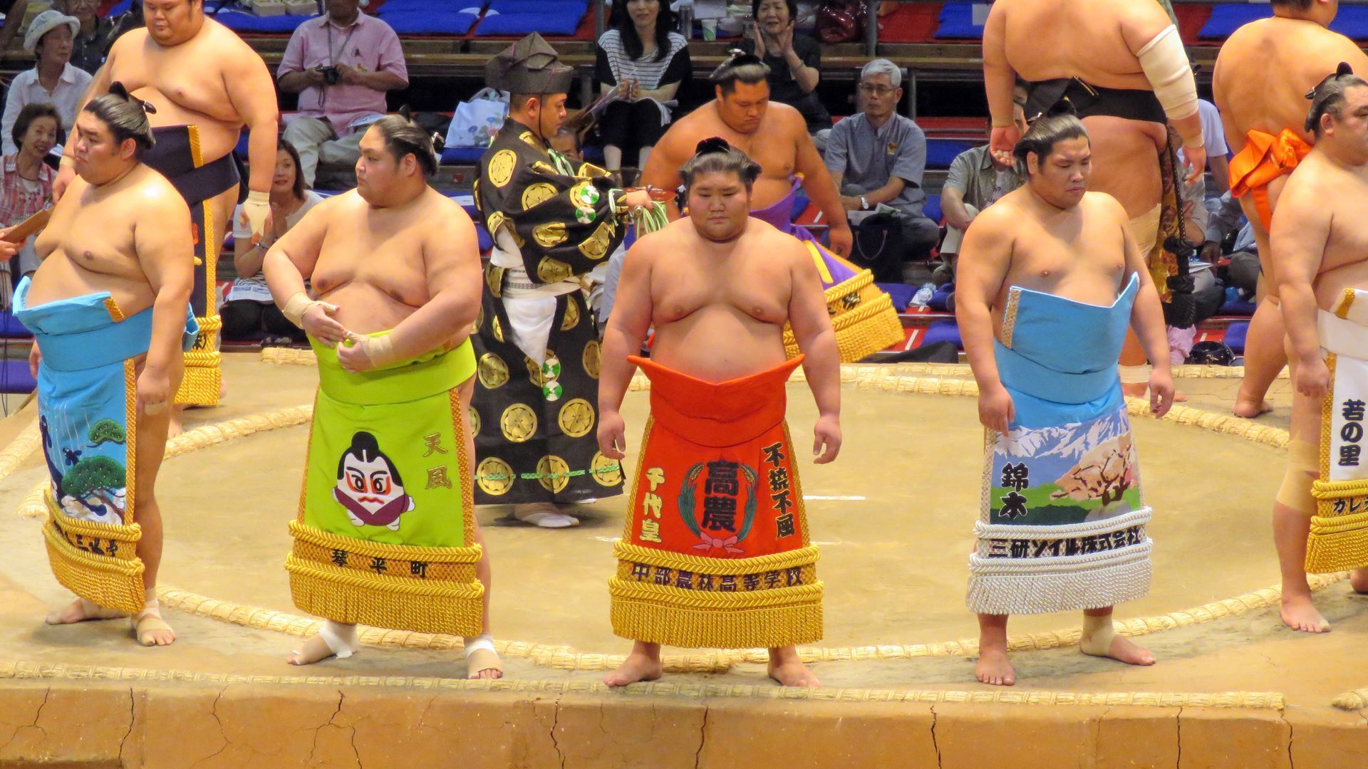 Sumo wrestlers in colorful aprons stand in a ring. Spectators in background.