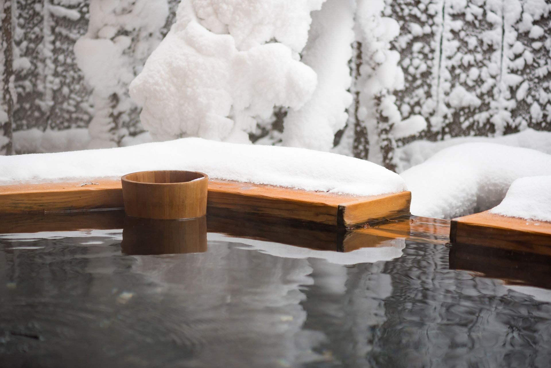 Wooden tub with hot water, snow-covered surroundings, and a bamboo cup.