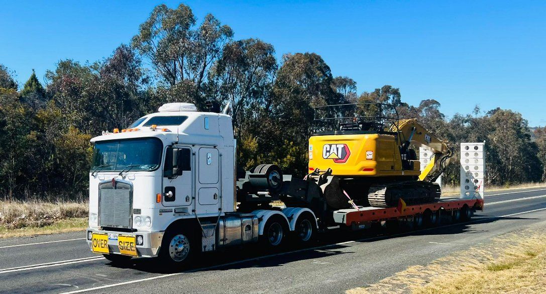 A White Semi Truck Is Driving Down a Road with A Yellow Excavator on The Back — RC Civil in Armidale, NSW