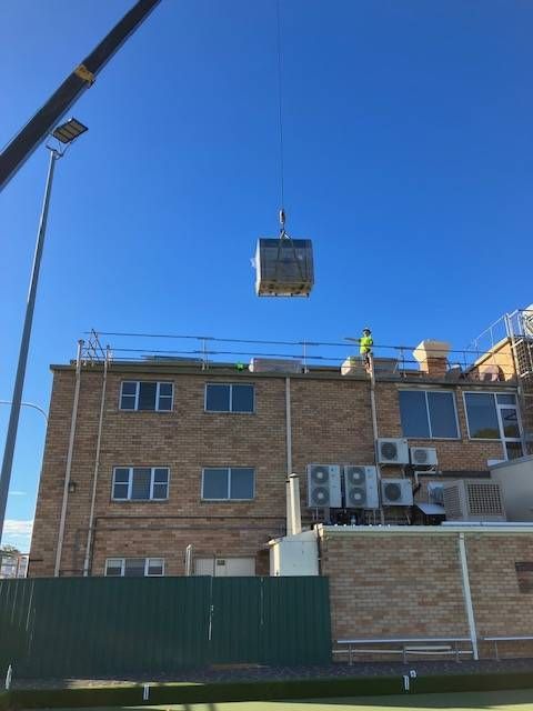 Crane lifting a small, metal box over a building. A worker is on the roof. Sunny day. — Todd Basham Electrical in Cootamundra, NSW