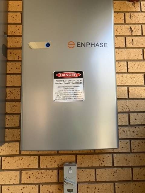 Silver Enphase solar panel box with warning label on a brick wall. — Todd Basham Electrical in Cootamundra, NSW
