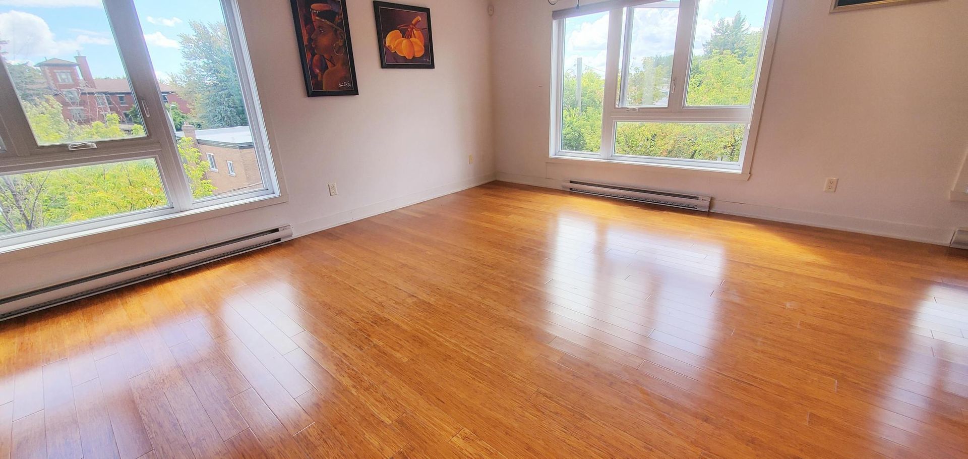 Chambre vide avec parquet poli, grandes fenêtres et œuvres d'art au mur.