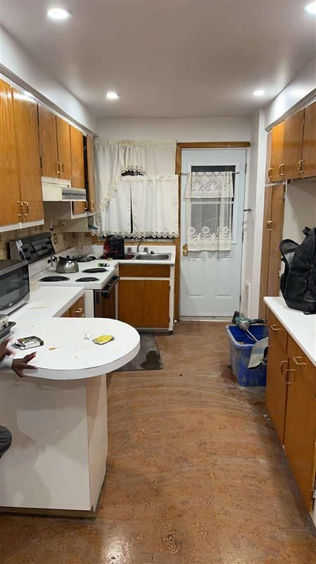 Cuisine avec armoires en bois clair, comptoirs blancs et sol marron. Une porte donne au fond de la pièce.