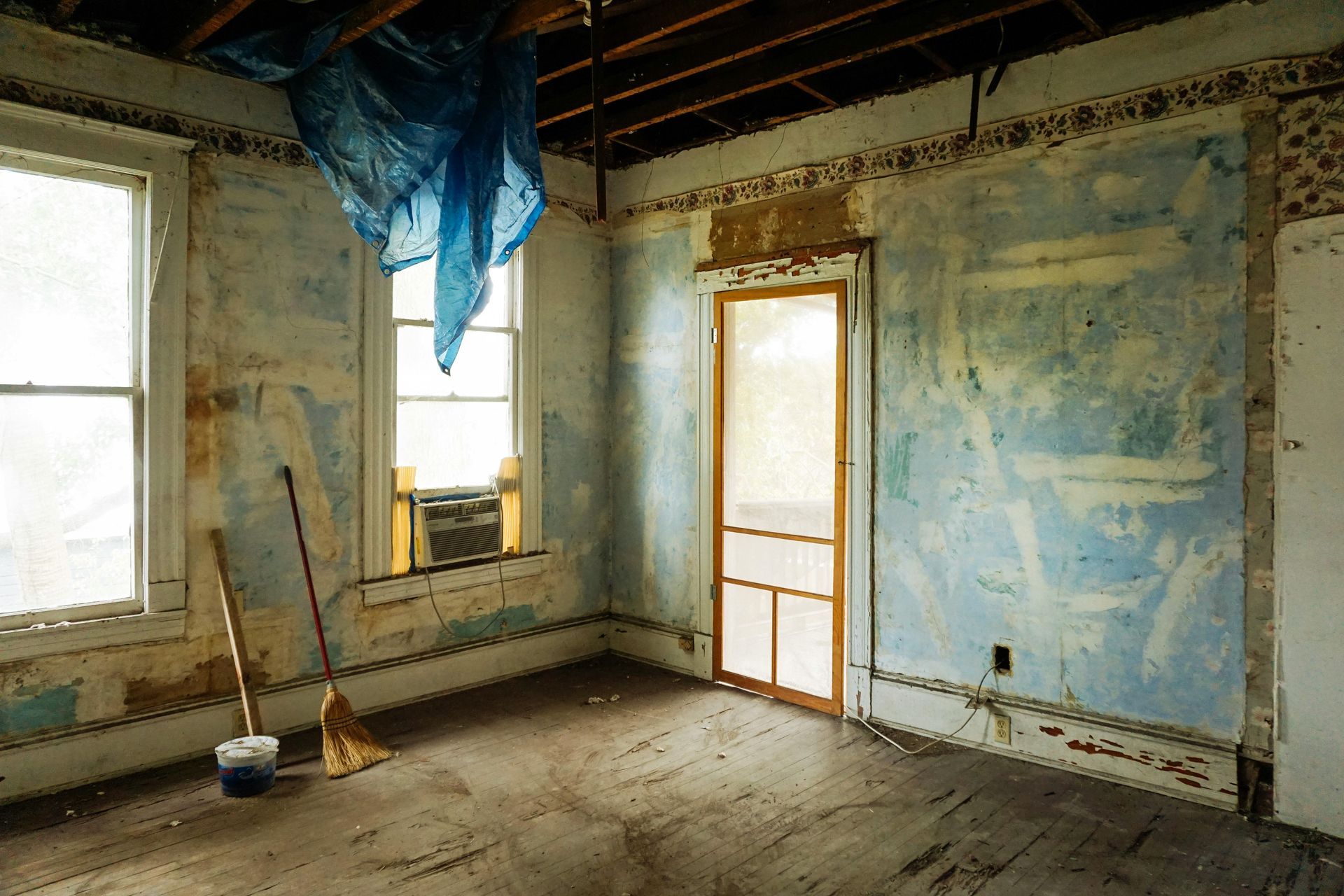 Dilapidated room with peeling blue walls, two windows, a doorway, and a broom leaning against a wall.