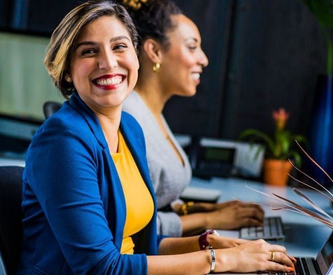 Two women are sitting at a desk using laptops and smiling.