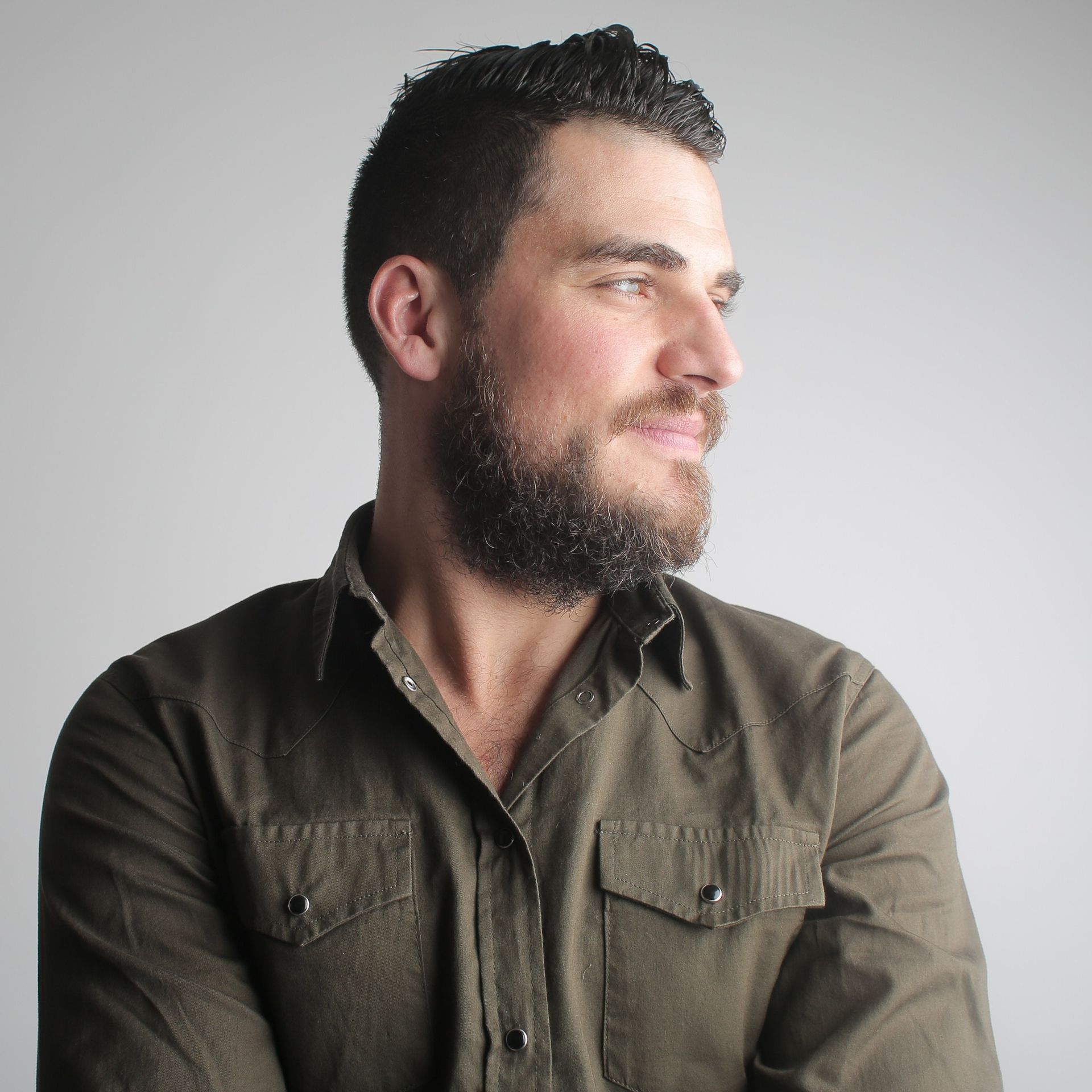 A man with a beard and short dark hair looks to the right, wearing a green button-up shirt against a light gray background.