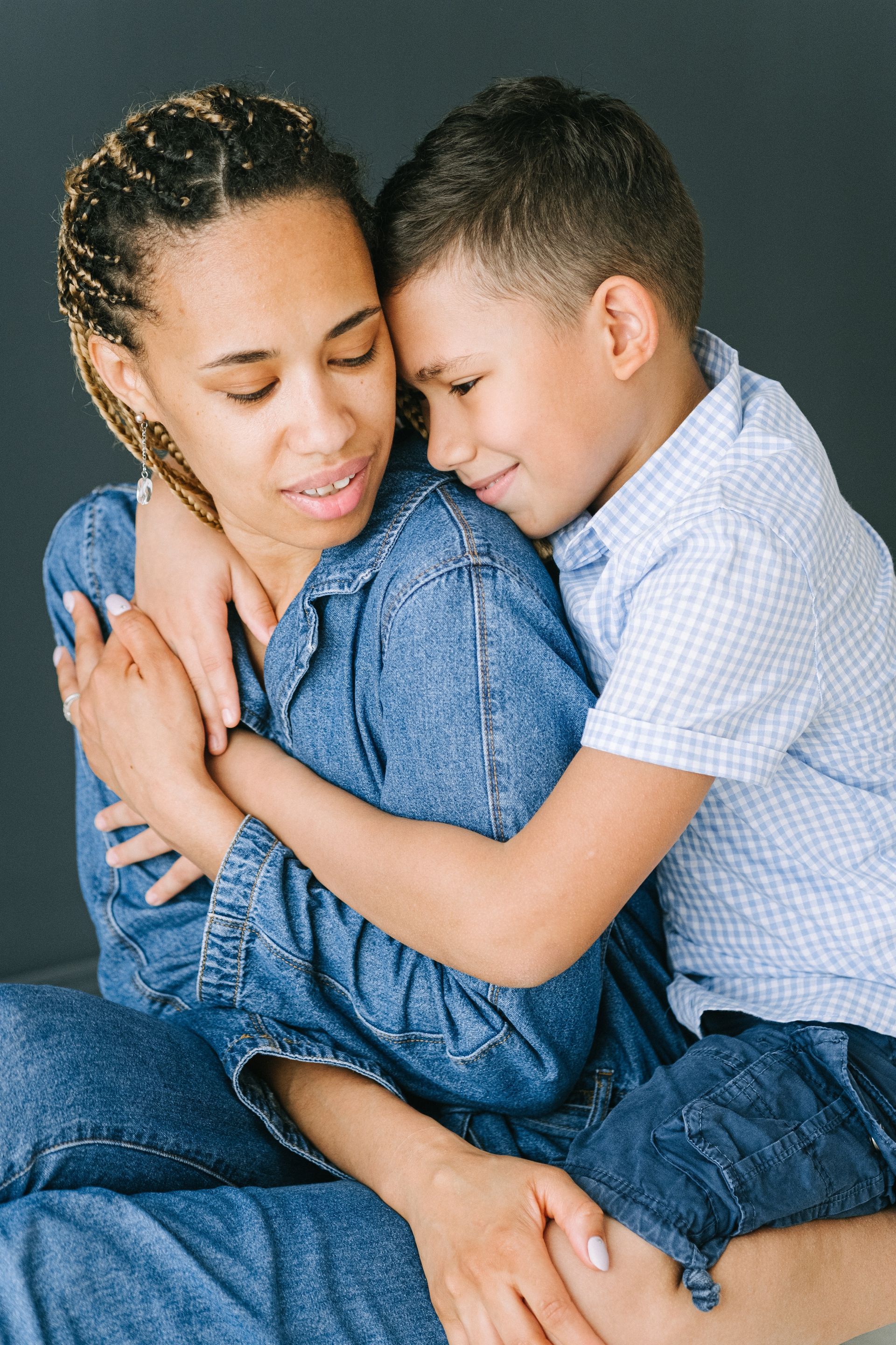 Woman in denim embraces a young boy, both smiling and close, against a dark background.