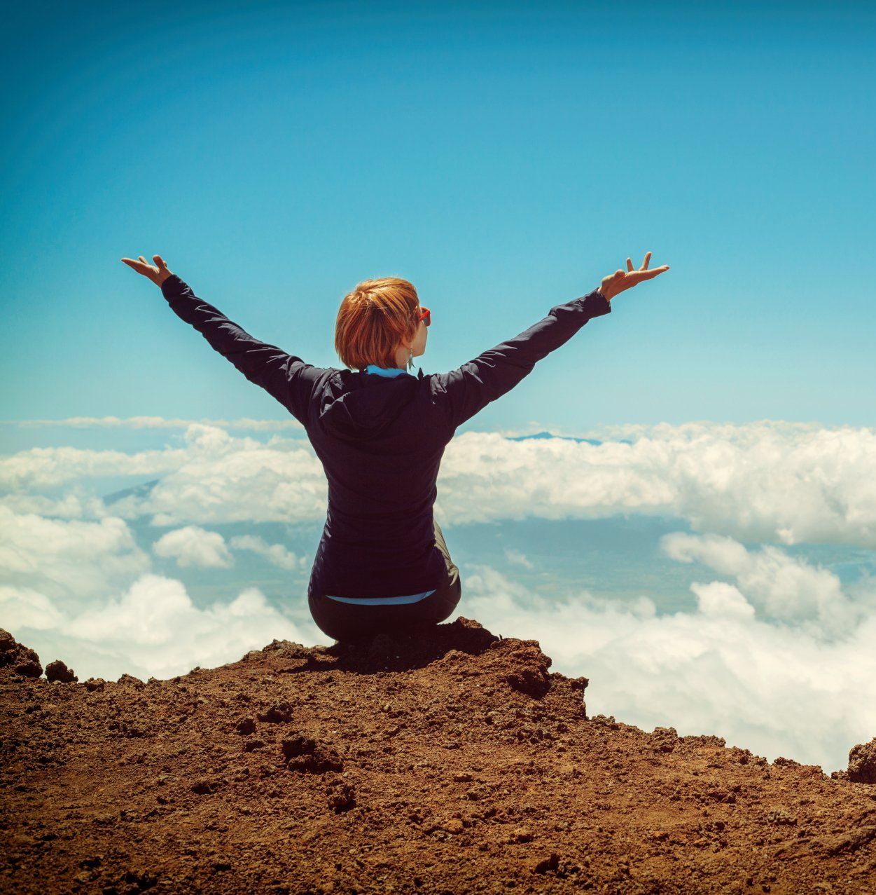 A woman is sitting on top of a hill with her arms outstretched