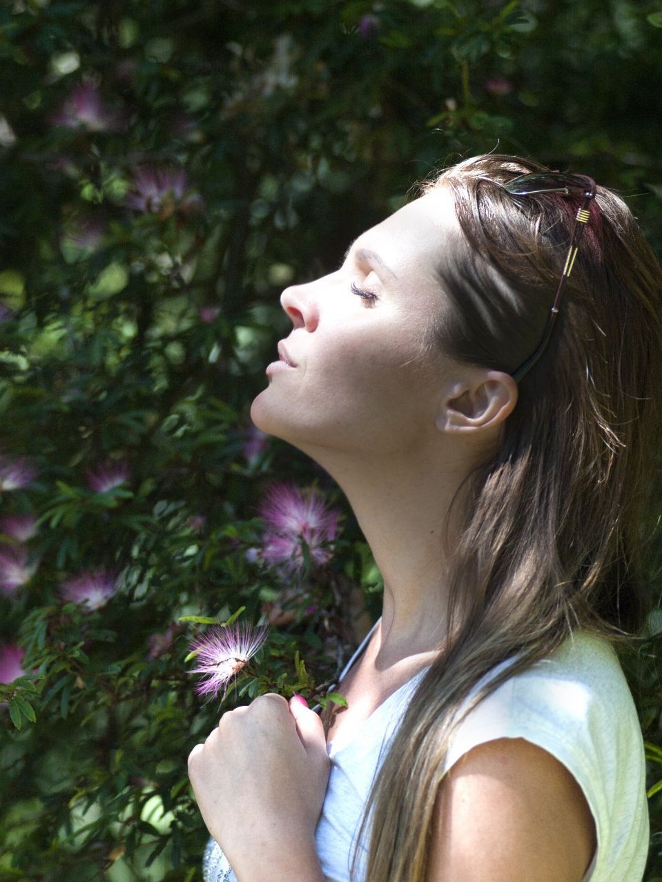 A woman is standing in front of a bush with her eyes closed.