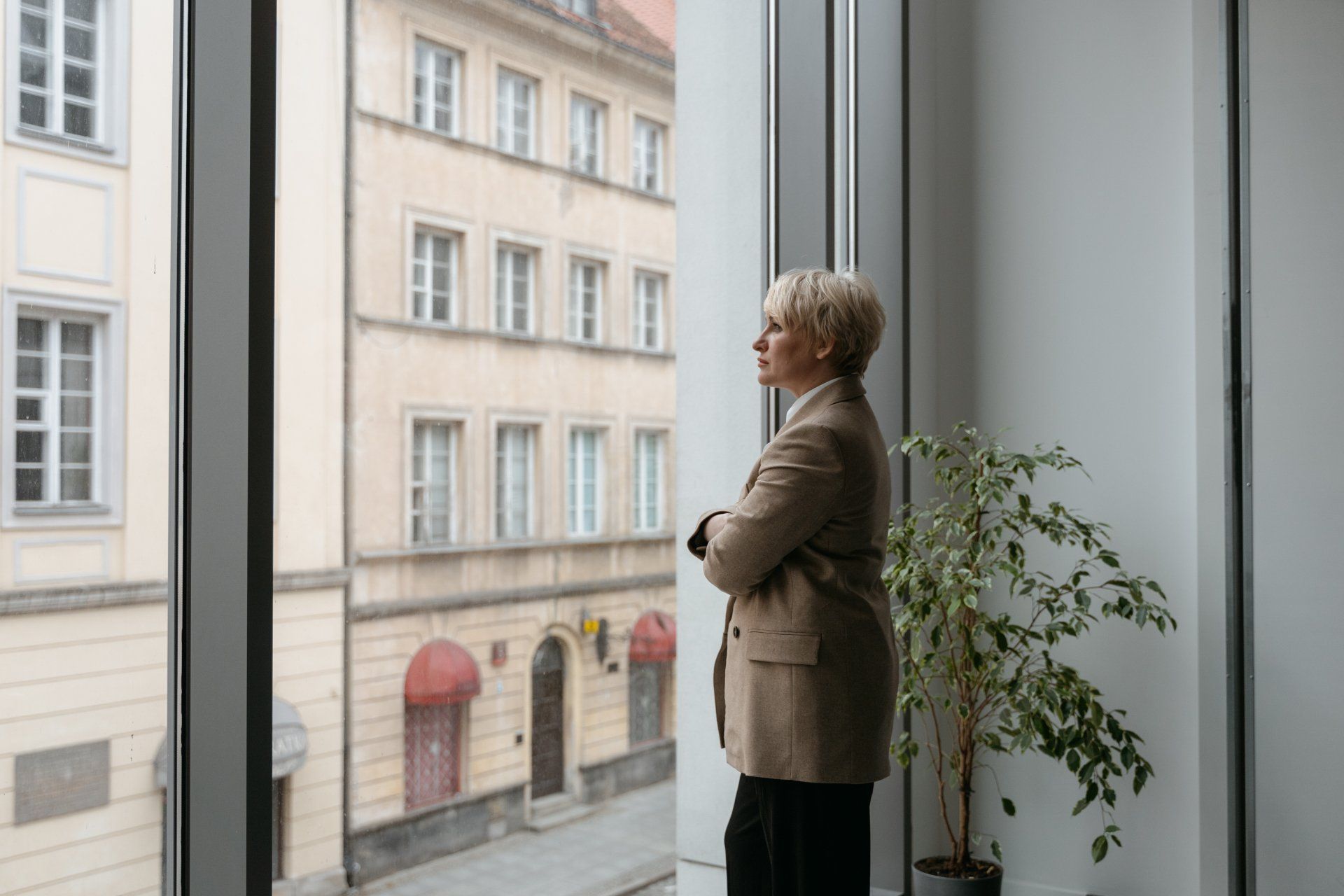 Woman in a tan blazer looking out a large window at an old building. She has her arms crossed.