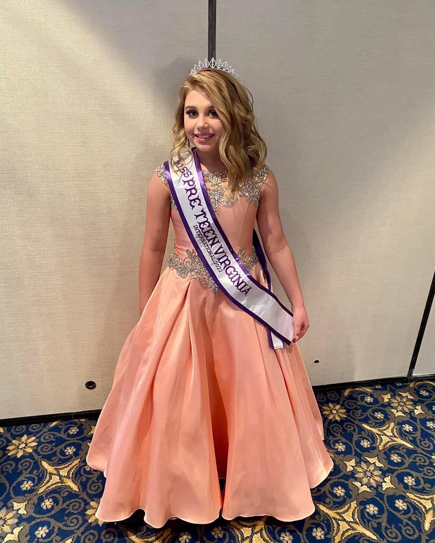A little girl in a dress and sash is standing in a room.