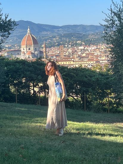 Woman in a dress, holding a tote bag, standing in a park with Florence, Italy, in the background.