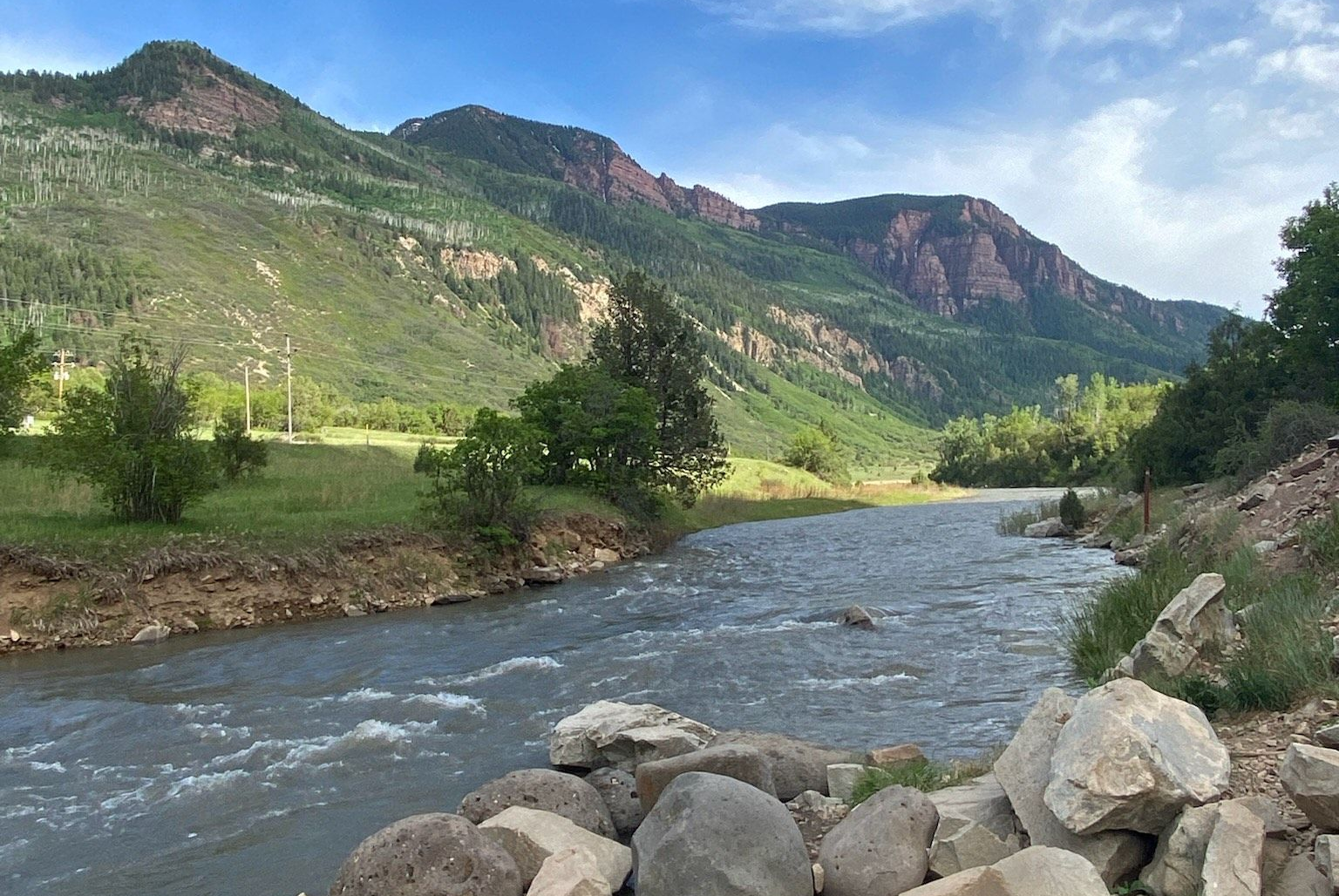 River flowing through a valley in Colorado with mountains in the background. Lush green vegetation and a blue sky.