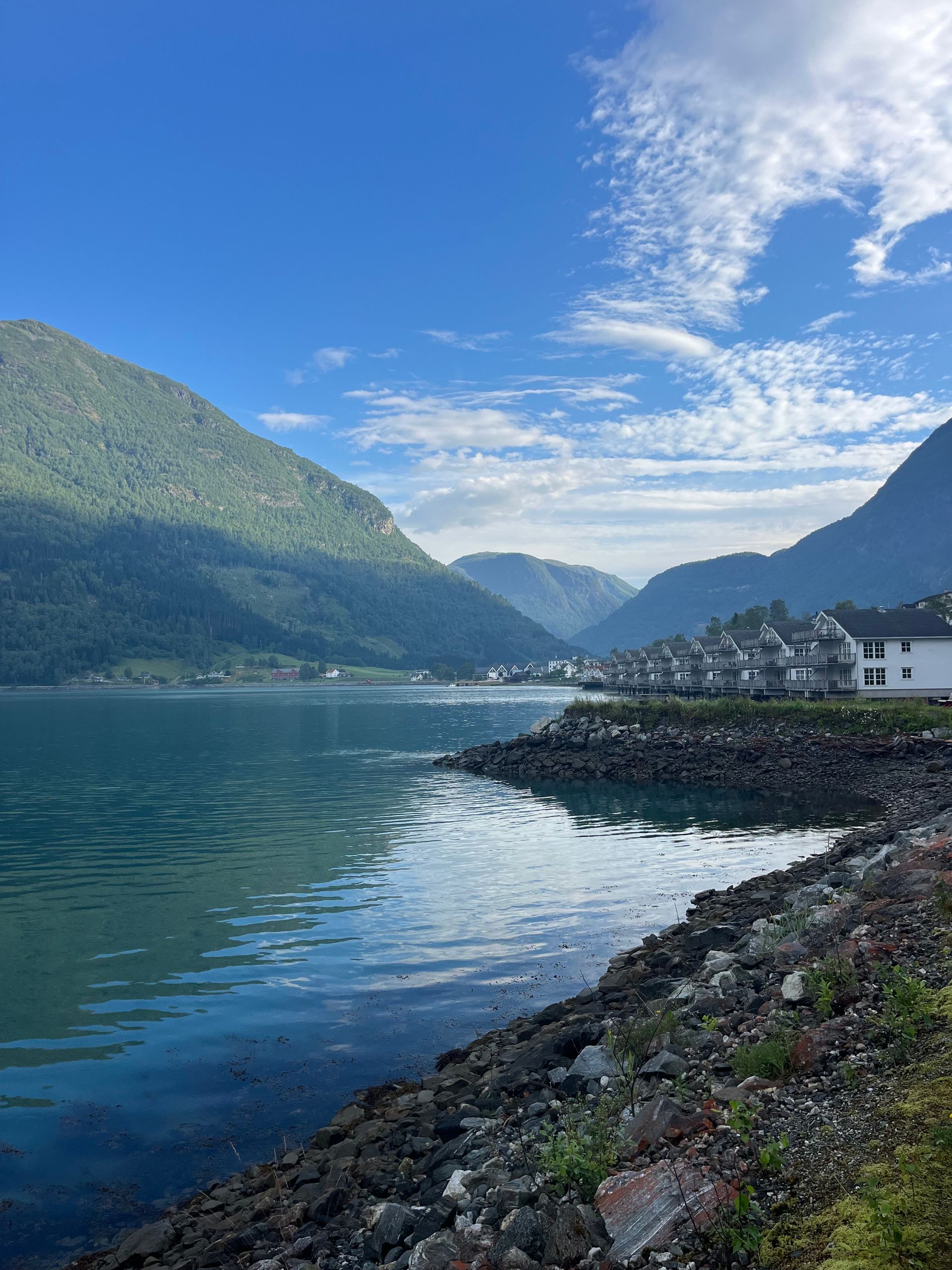 Serene coastal view: Teal water, rocky shore, lush mountains, and a village under a blue, cloudy sky.