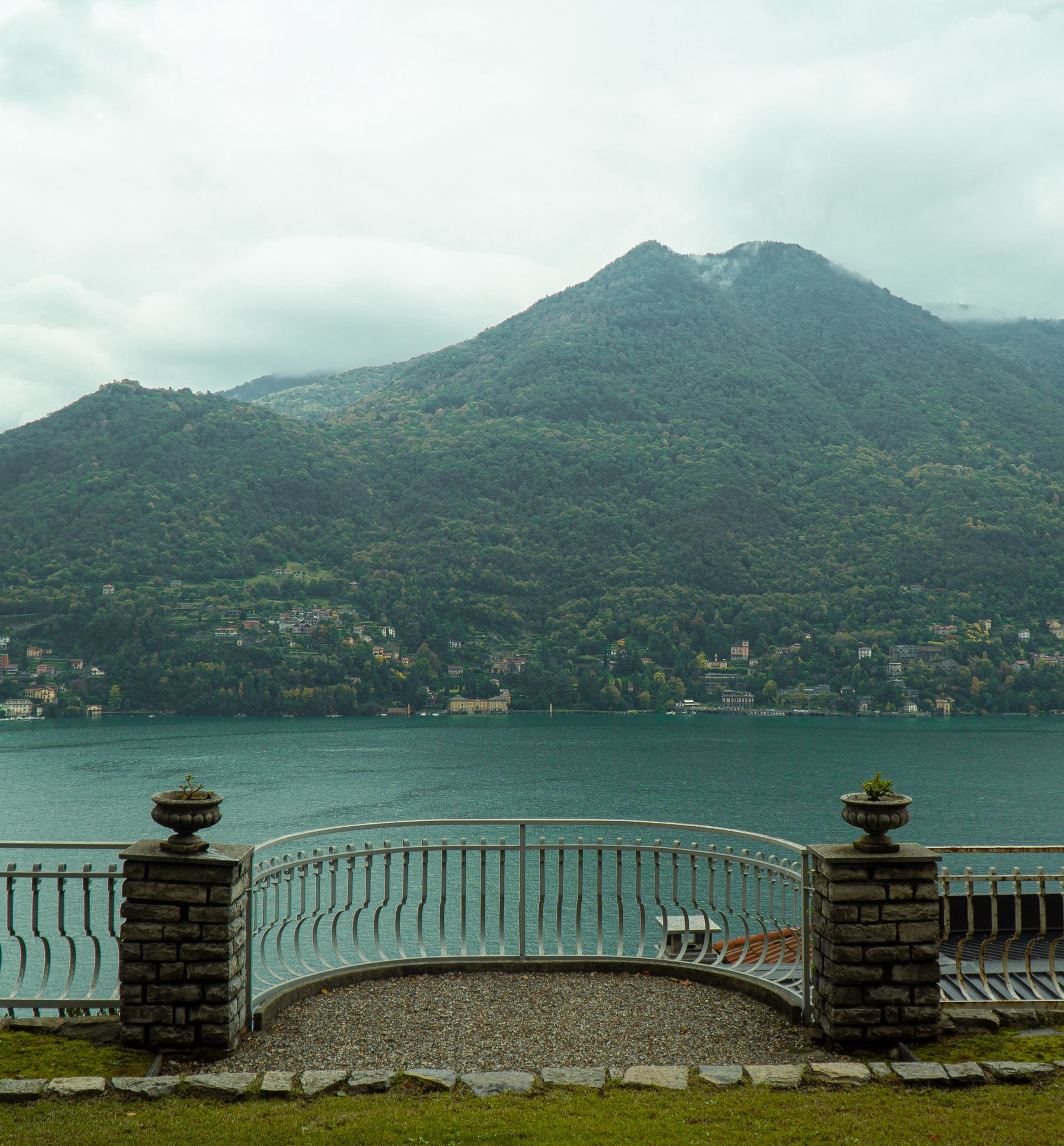 Lake Como, Italy view with mountain backdrop. Balcony with stone pillars, metal railing, and blue water. Overcast sky.