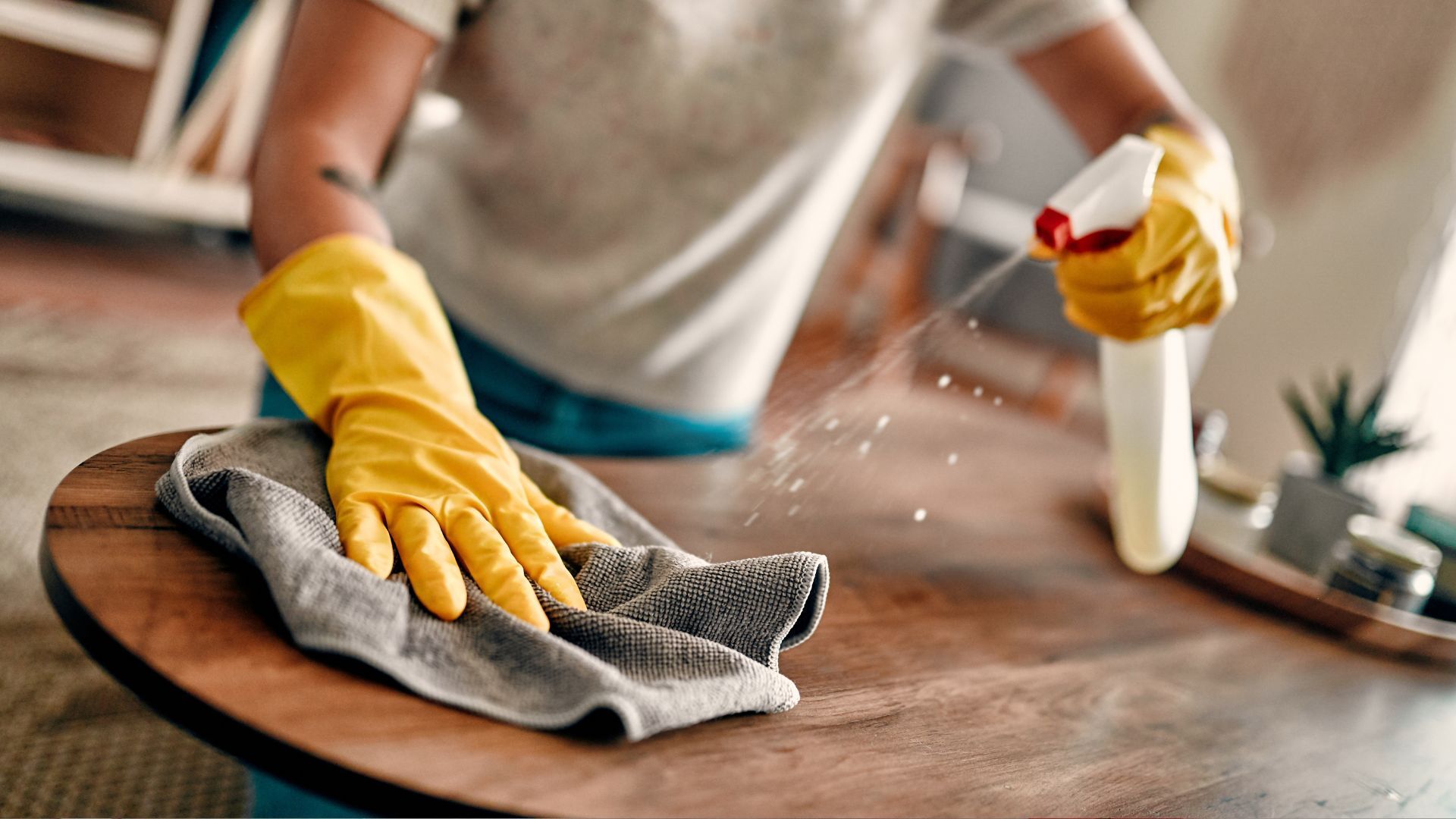 A person wearing yellow gloves is cleaning a wooden table with a cloth and spray bottle.