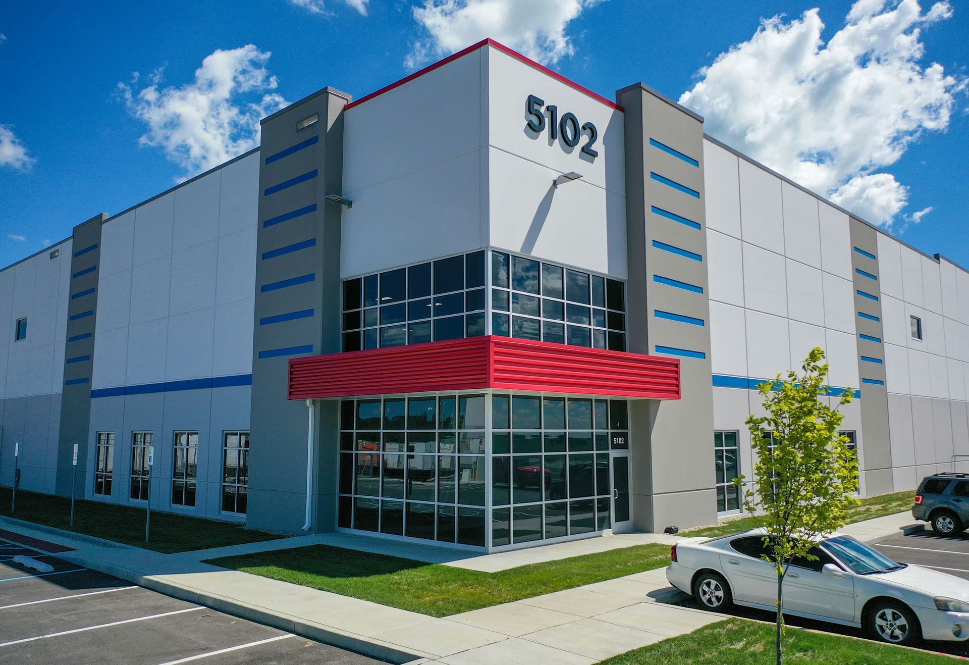 Shopping center with various storefronts under a clear blue sky, cars parked in front.