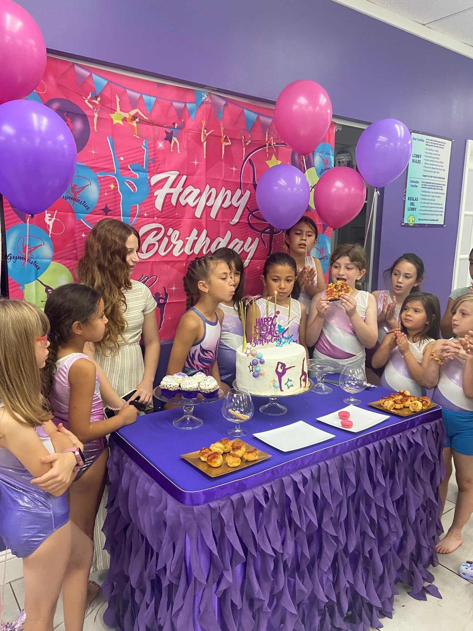 Un grupo de niños están de pie alrededor de una mesa con un pastel de cumpleaños y globos.