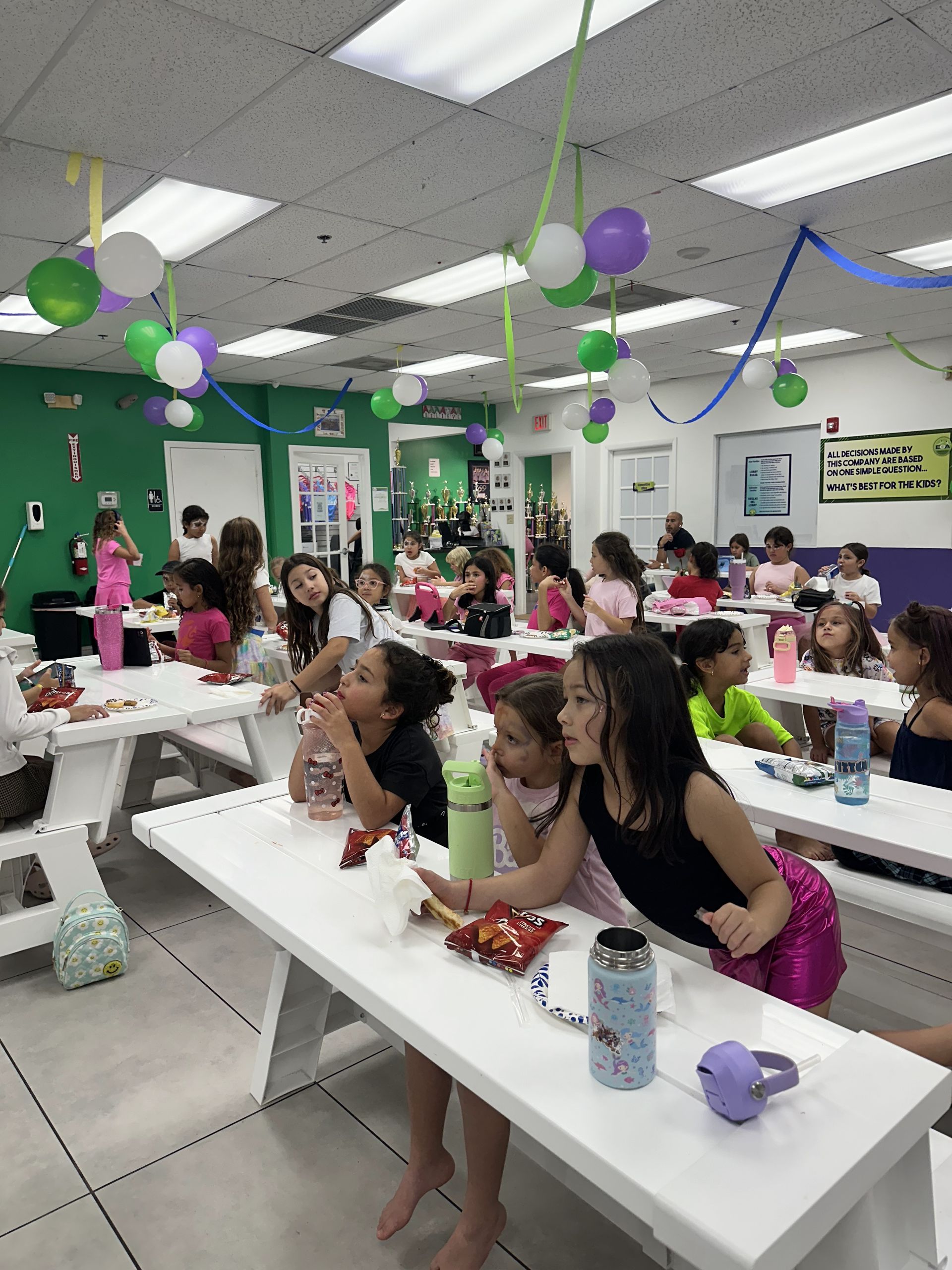 Un grupo de niños están sentados en mesas de picnic en una habitación decorada con globos.