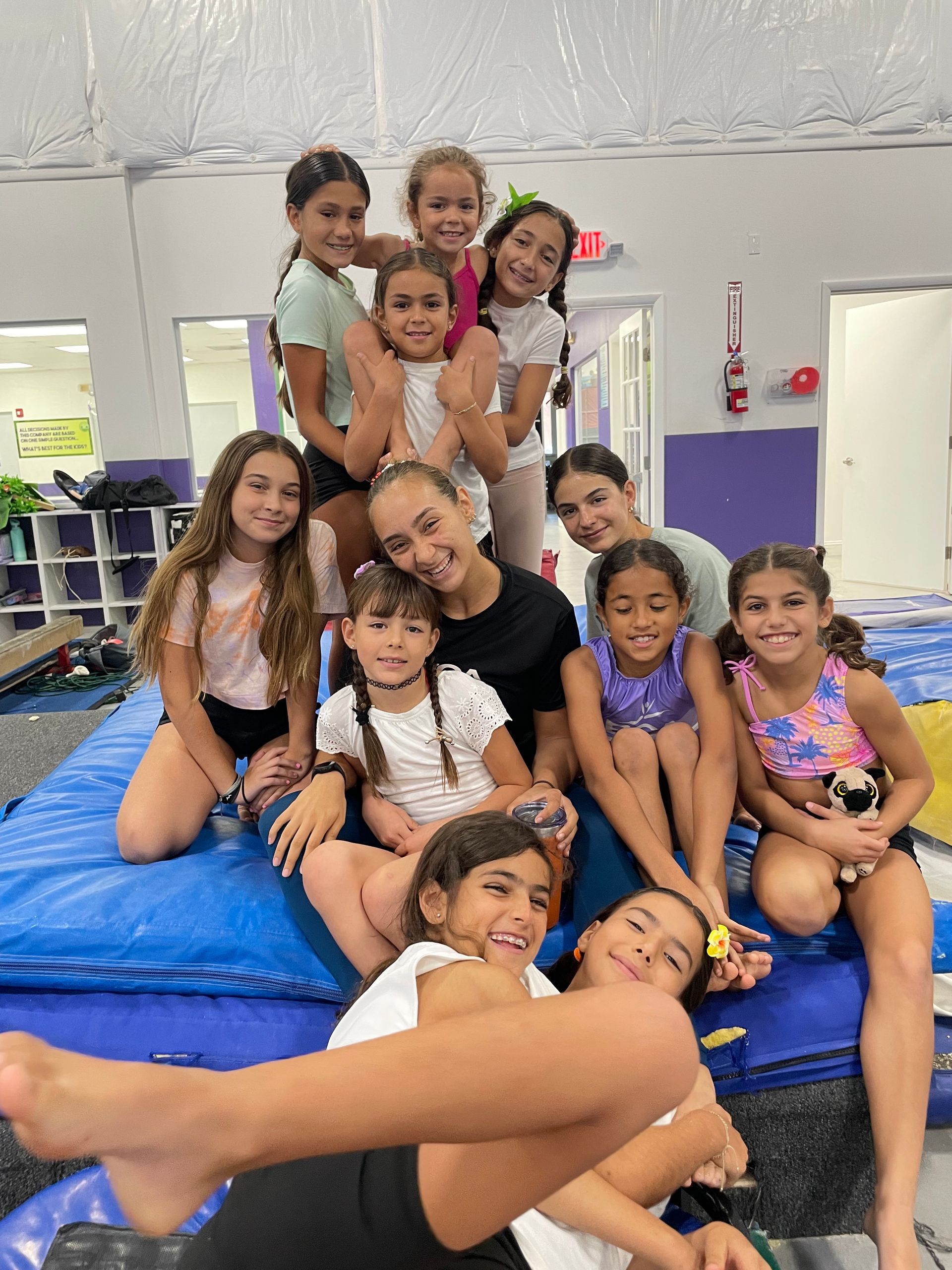 Un grupo de chicas jóvenes están sentadas encima de una colchoneta azul en un gimnasio.