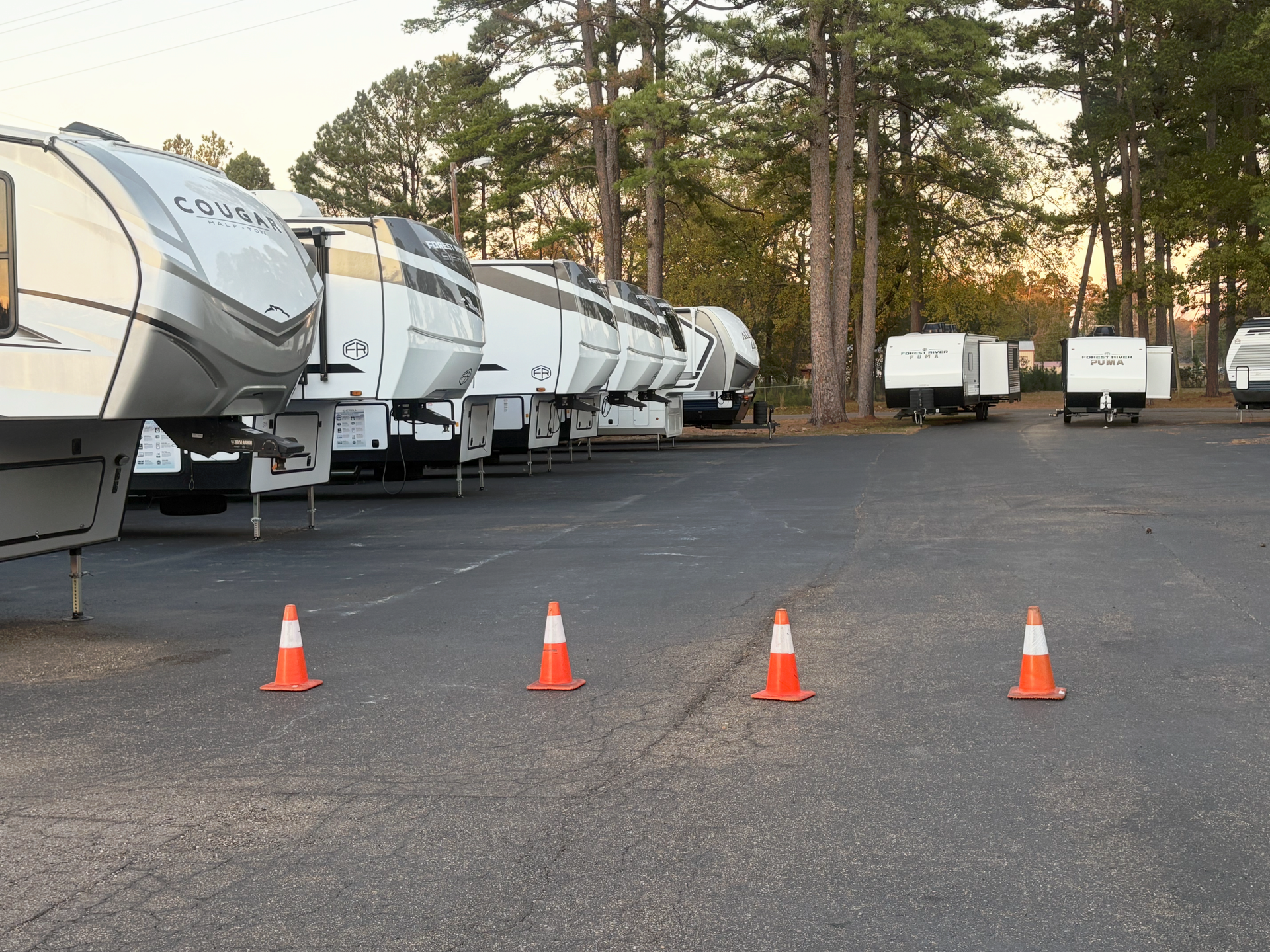 Row of white RVs parked on asphalt, with orange traffic cones in the foreground. Trees in the background.