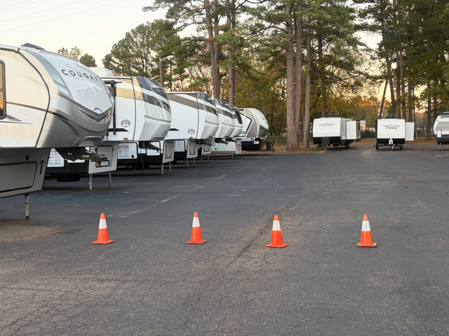 Row of white RVs parked on asphalt, with orange traffic cones in the foreground. Trees in the background.