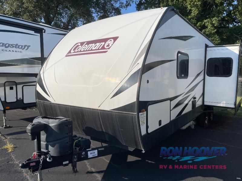 White Coleman travel trailer with open door, parked outdoors.