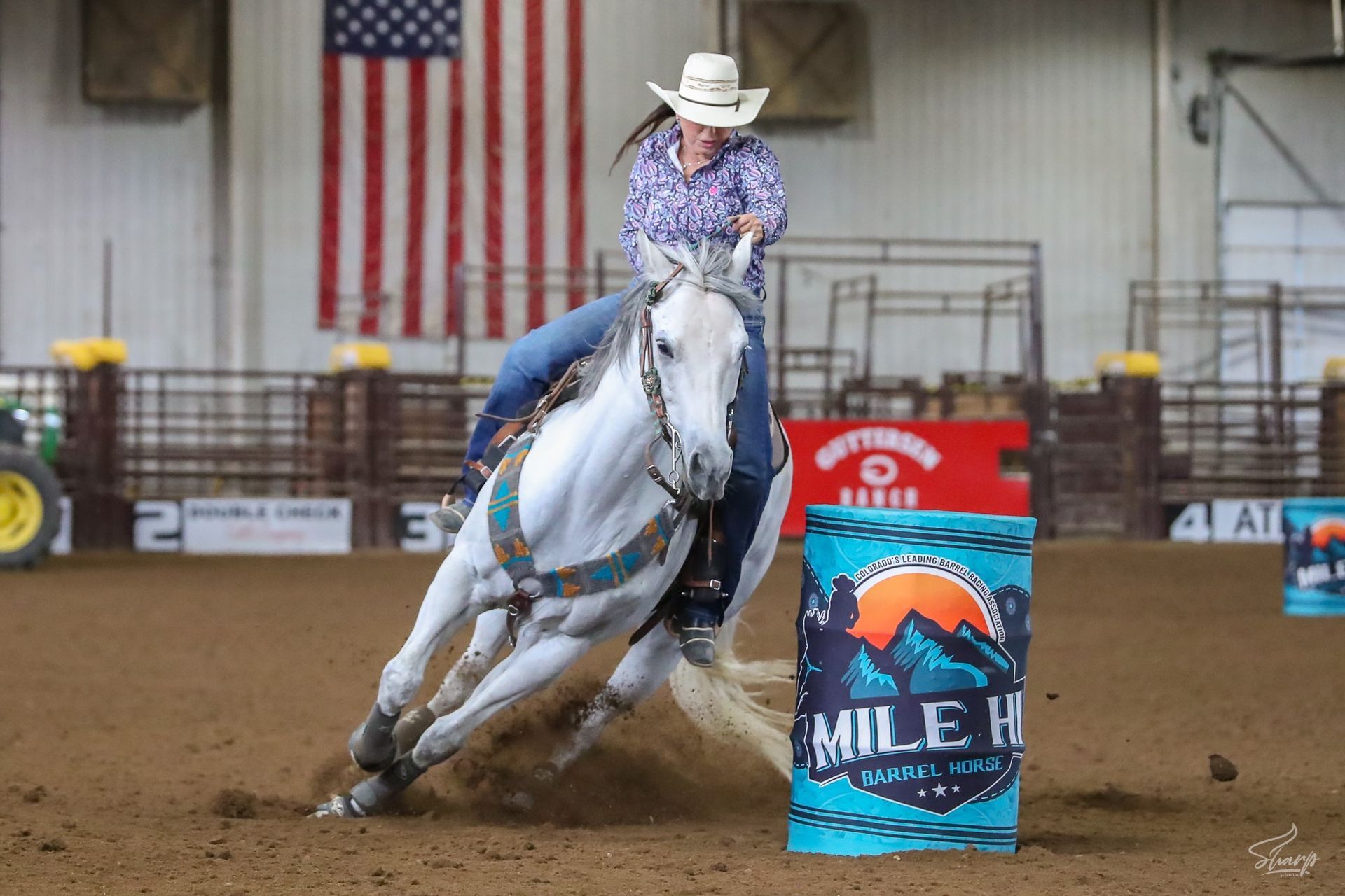 A woman is riding a white horse around a barrel in a rodeo arena.