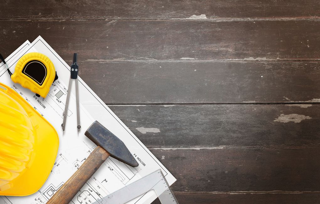 A yellow hard hat, tape measure, compass, hammer, and ruler on architectural blueprints over a dark wooden surface.