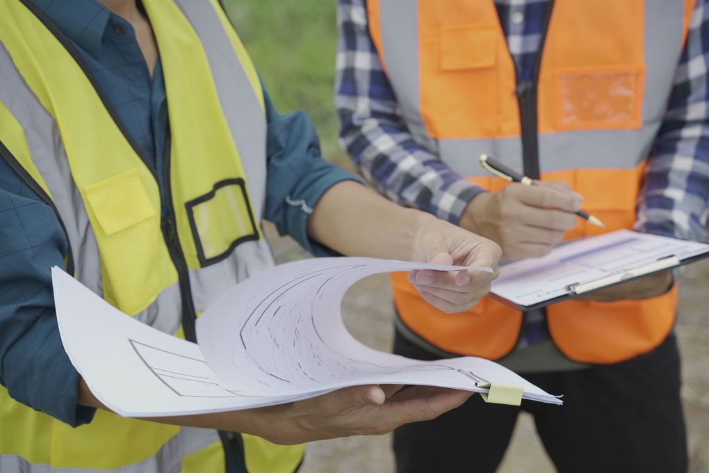 Two construction workers in safety vests review plans and take notes on a clipboard outdoors.
