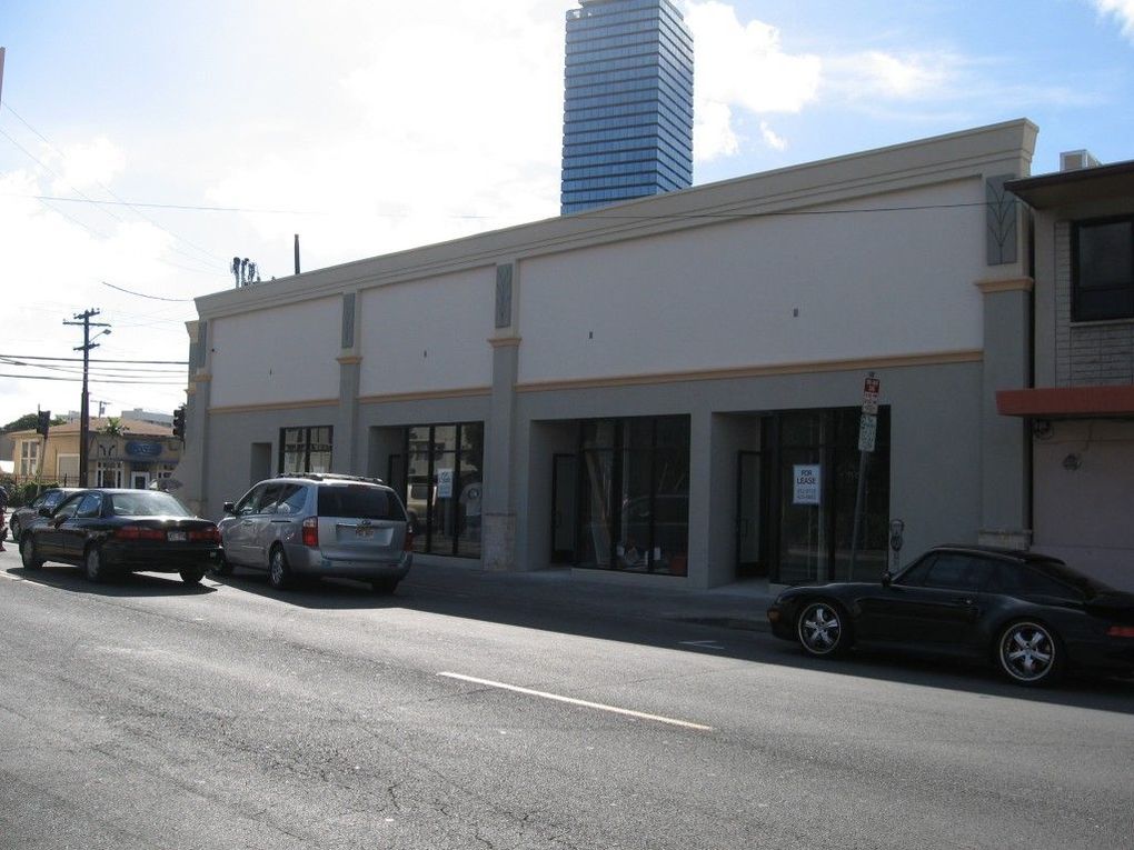 A beige commercial building with glass storefronts along a street, with parked cars and a tall skyscraper in the background.