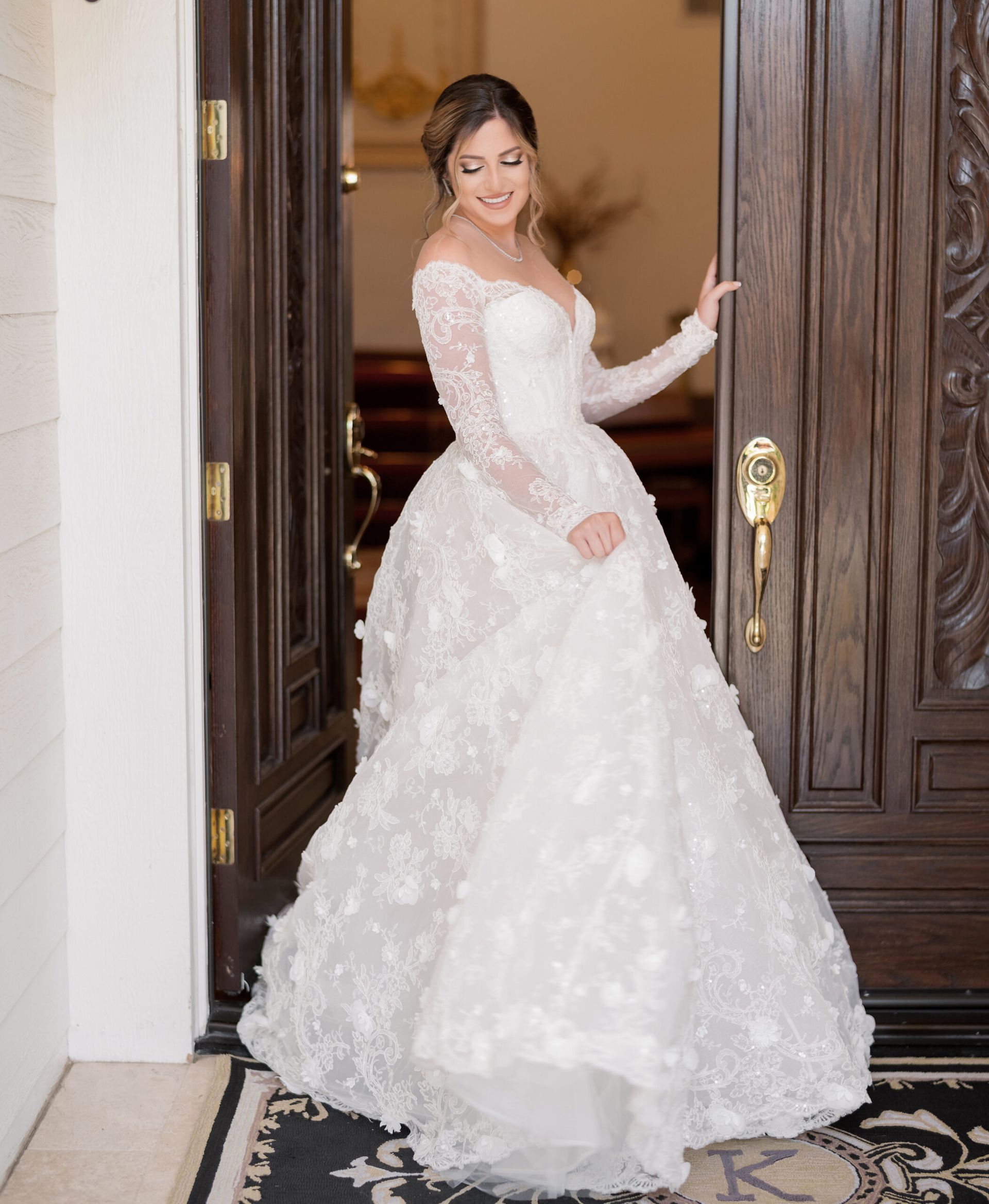 Bride in white lace wedding dress stands at open wooden door, smiling.