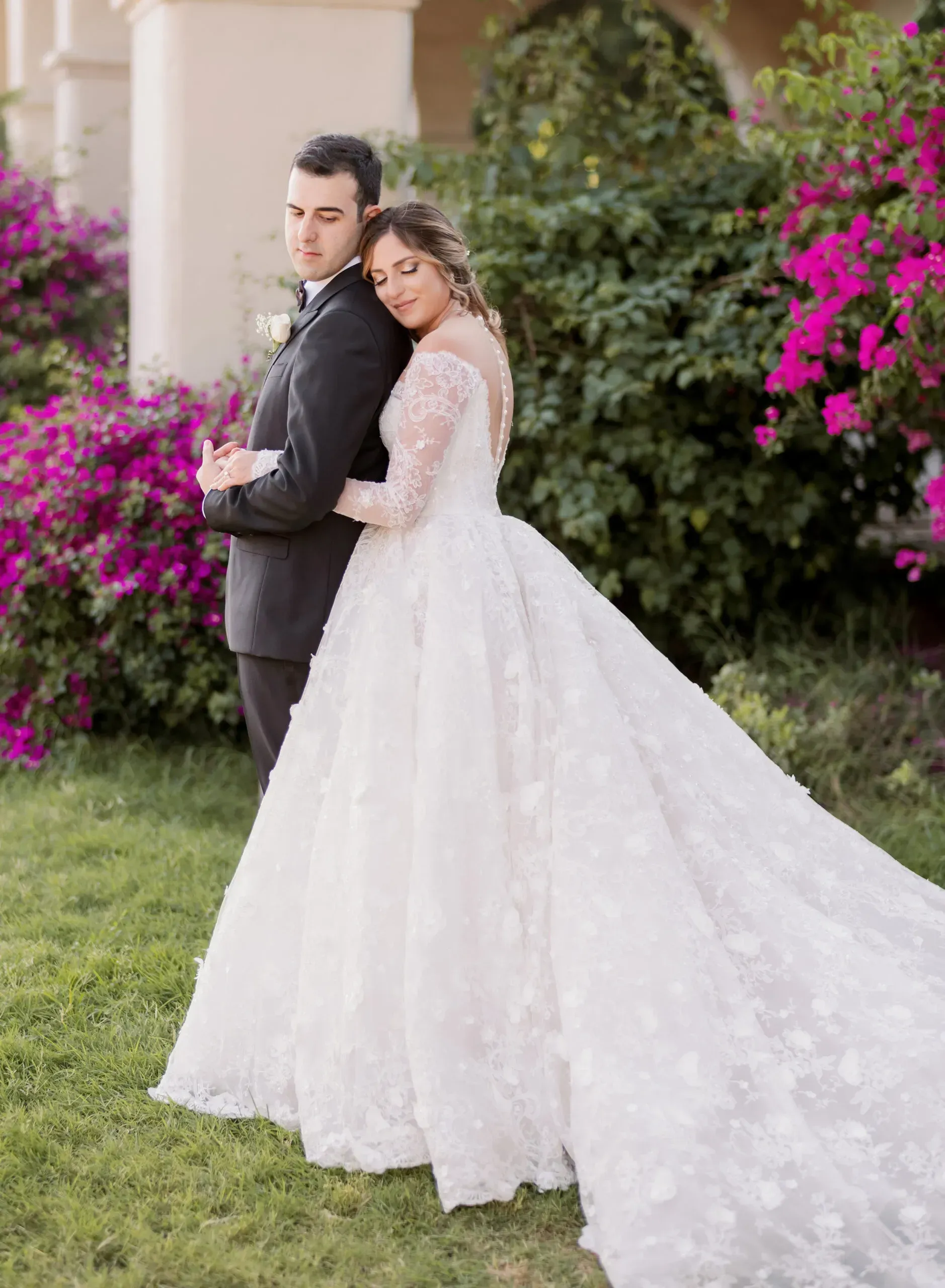 Bride in white gown leans on groom in black suit outdoors with flowers.