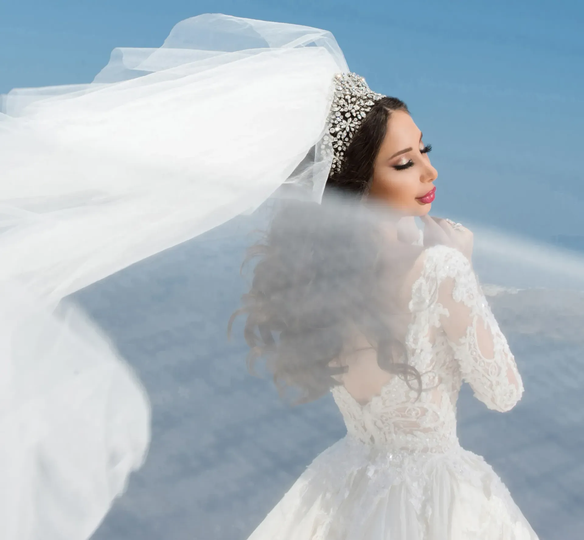 Bride in white lace dress and veil, smiling, with a jeweled headpiece, outdoors.