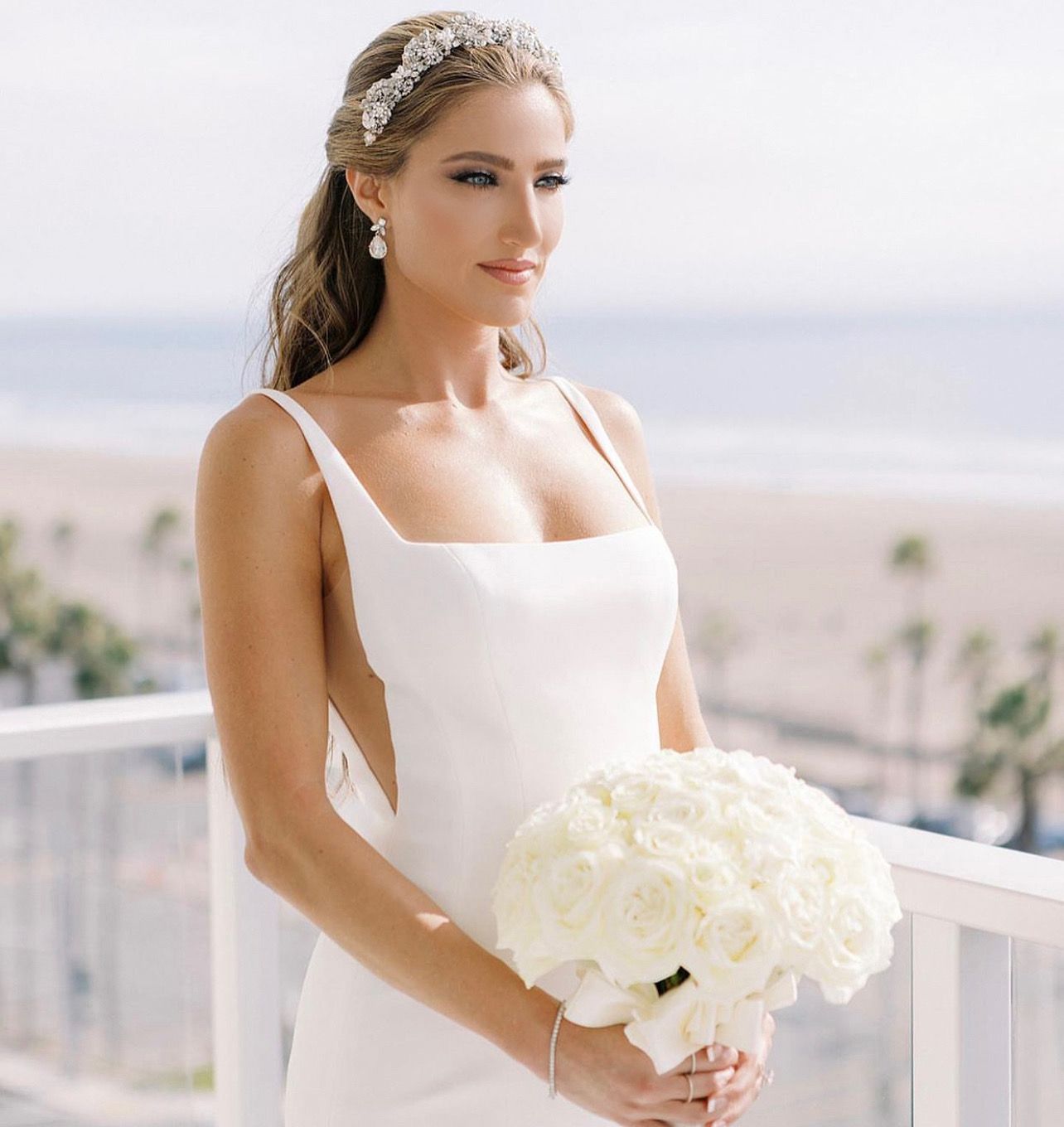 Bride in a white dress holds white roses, wearing a tiara, looking towards the ocean on a balcony.