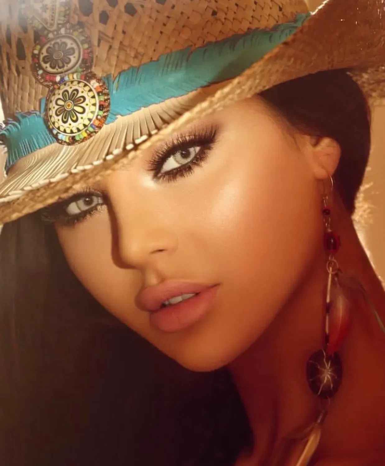 Woman with light eyes, wearing a decorated straw hat with turquoise accents and dangling earrings, bathed in warm light.