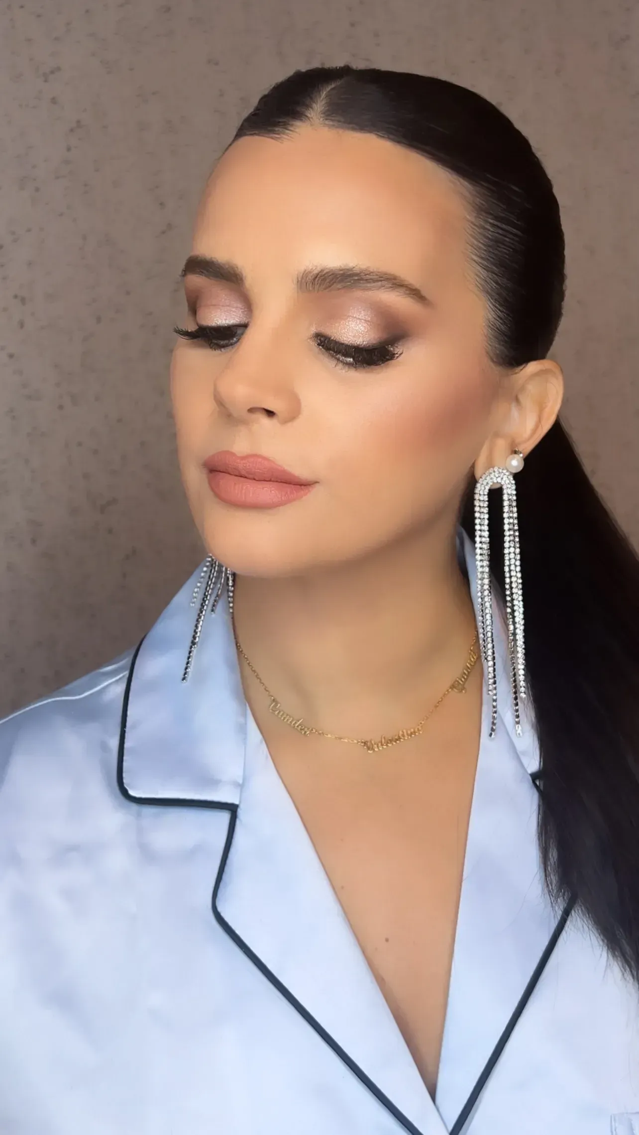 Woman with sleek ponytail, glamorous makeup, and dangling silver earrings, wearing a light blue pajama top.