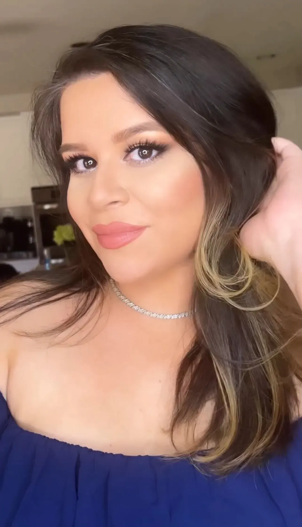 Woman with dark hair, wearing blue off-shoulder top, smiles at the camera, hand in hair.