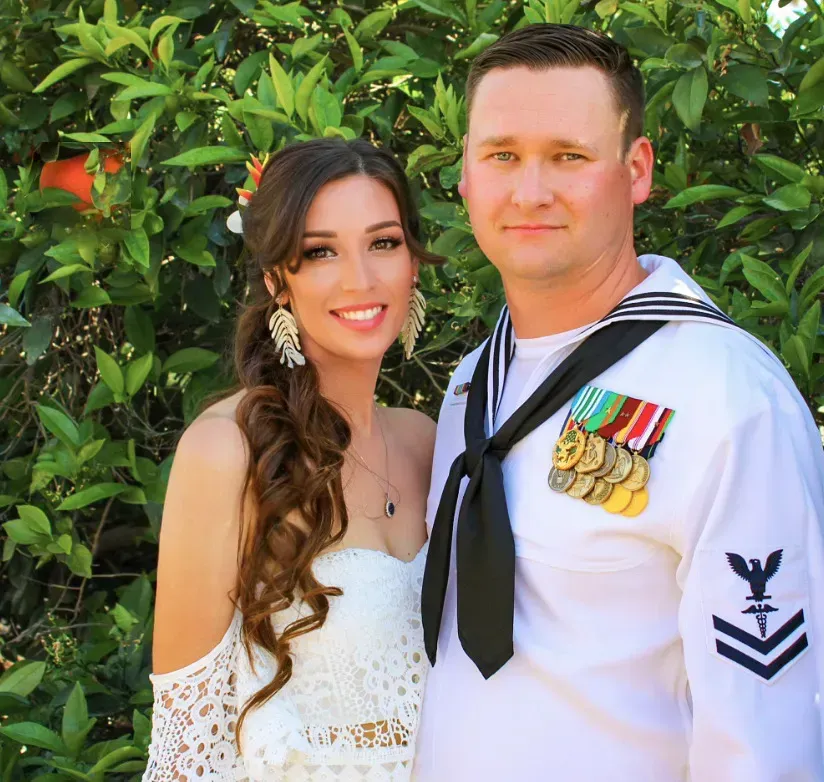 A smiling woman in white dress and a man in navy uniform pose outdoors.