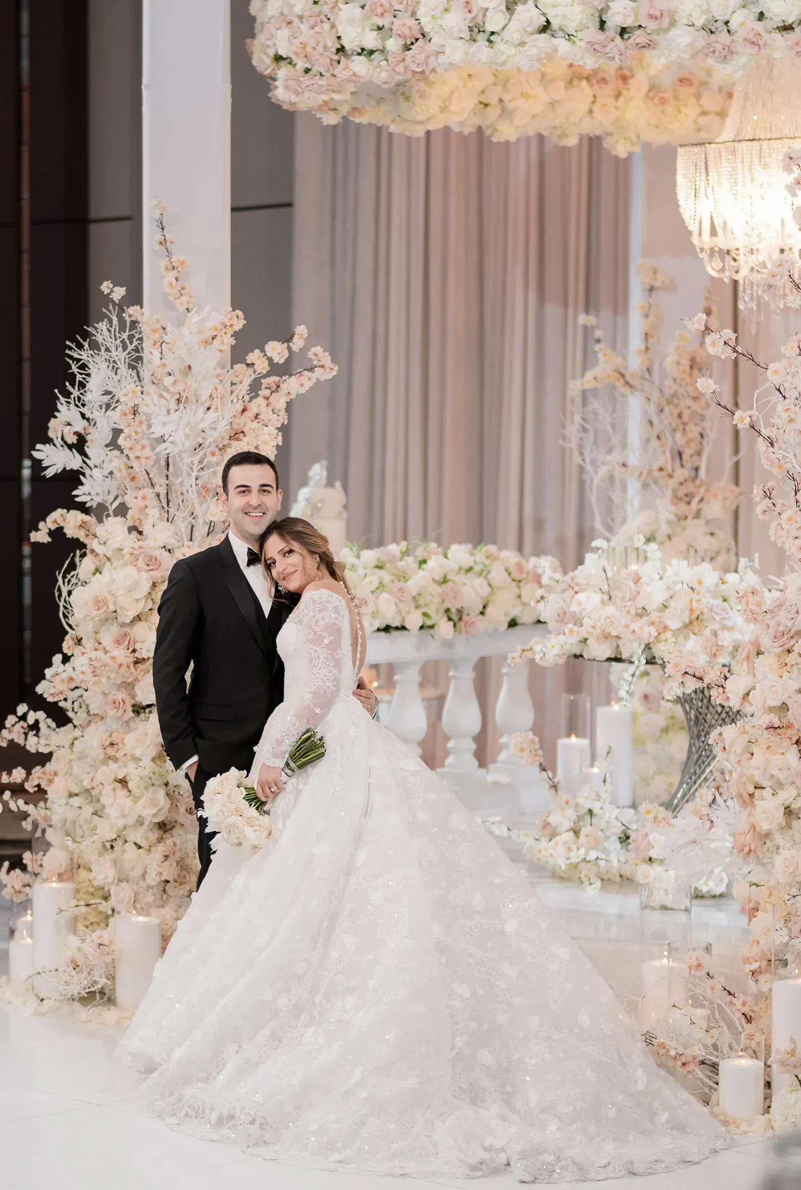 Bride and groom pose for wedding photo. Woman in white gown, man in black suit. Floral arch in background.