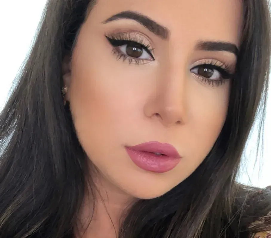 Woman with brown eyes, dark hair, and full makeup, including winged eyeliner and pink lipstick.