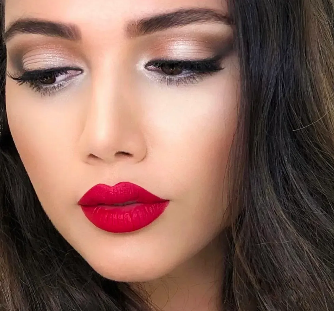 Woman with dark hair, wearing red lipstick and dramatic eye makeup, looking down.