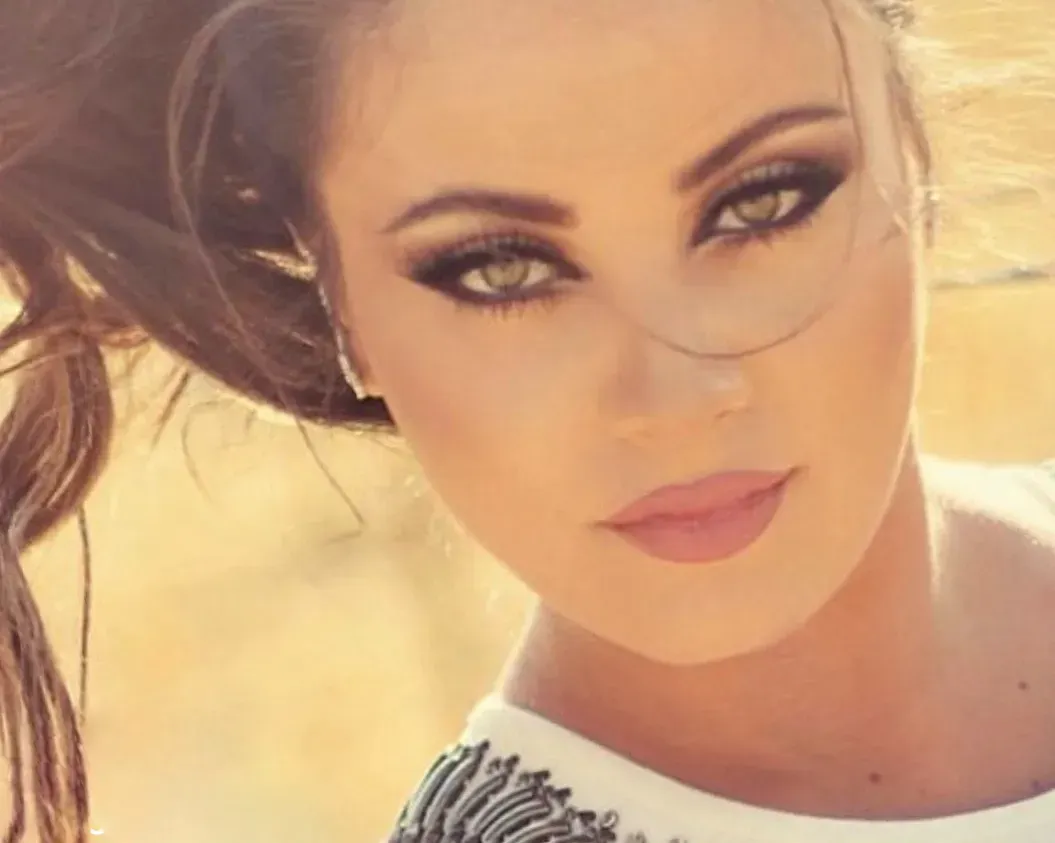 Woman with smoky eye makeup gazes toward viewer; brown hair blowing in the wind; sunlit background.