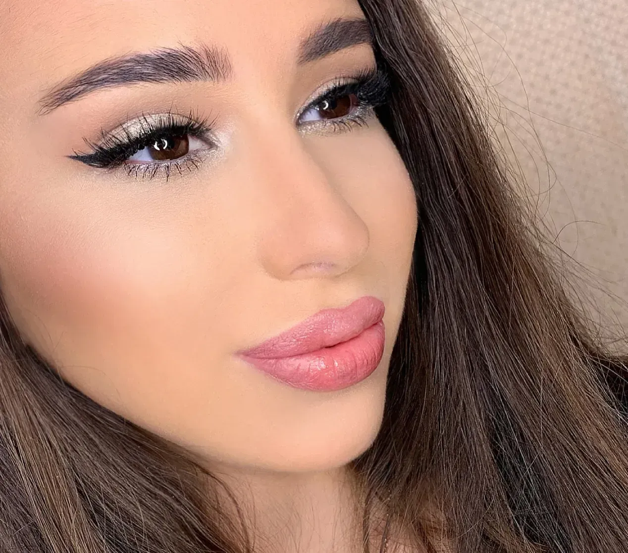 Woman with brown hair, wearing makeup: winged eyeliner, pink lipstick, and highlighted skin.