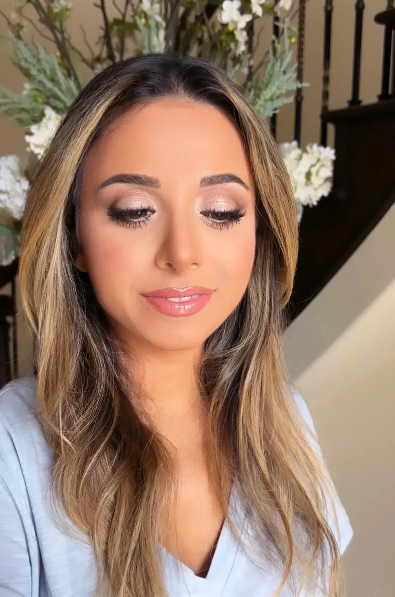 Woman with warm-toned makeup, light brown hair, and soft pink lips; in front of a staircase.