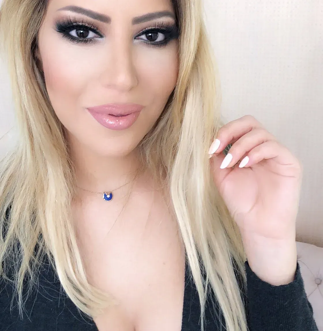 Blonde woman with dramatic eye makeup and a light pink lip, wearing a black top and gold necklace.