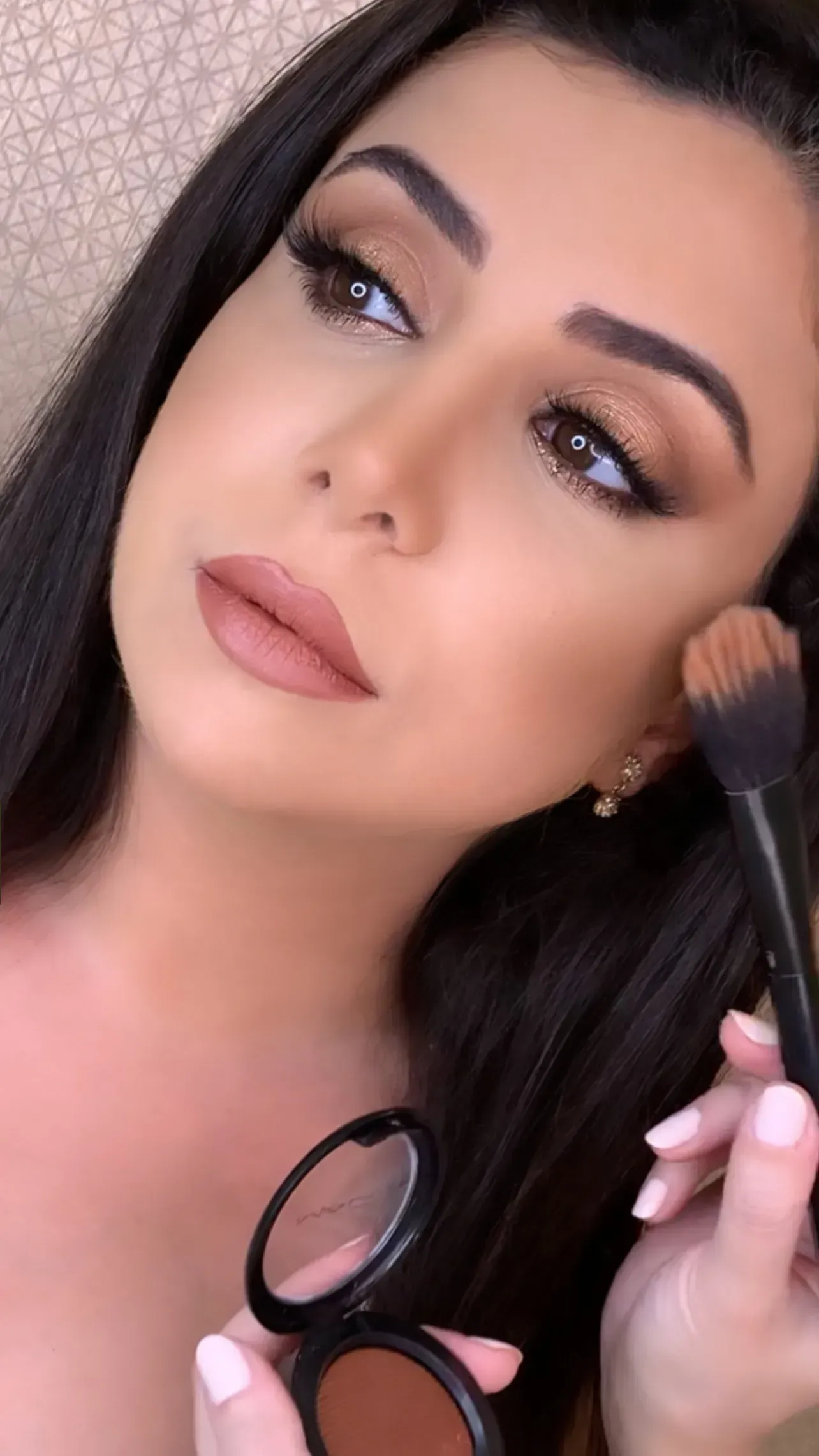 Woman with brown hair and makeup applies bronzer with a brush.