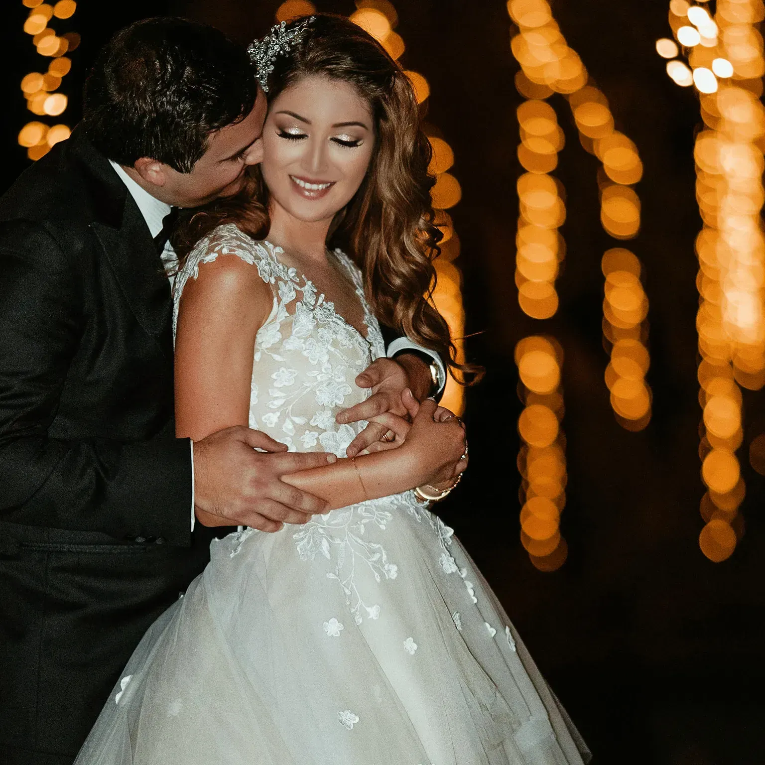 Groom kissing bride's cheek. They are embraced, in formal attire, with string lights in background.