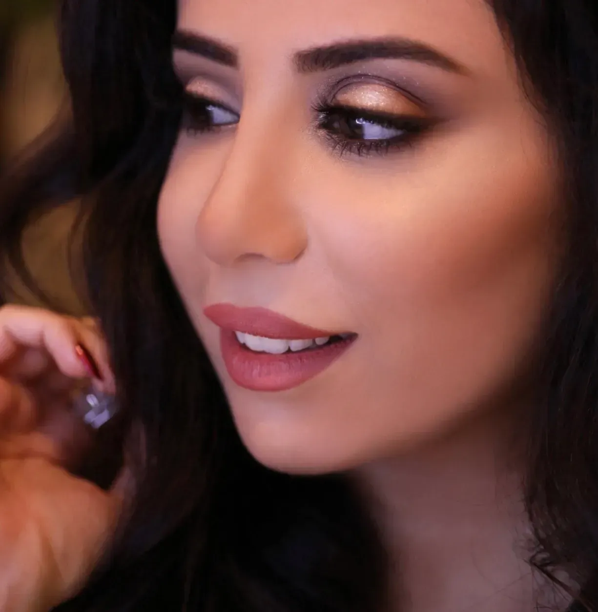 Woman with dark hair and glamorous makeup, looking slightly off-camera. Gold eyeshadow, pink lipstick, and a soft focus background.