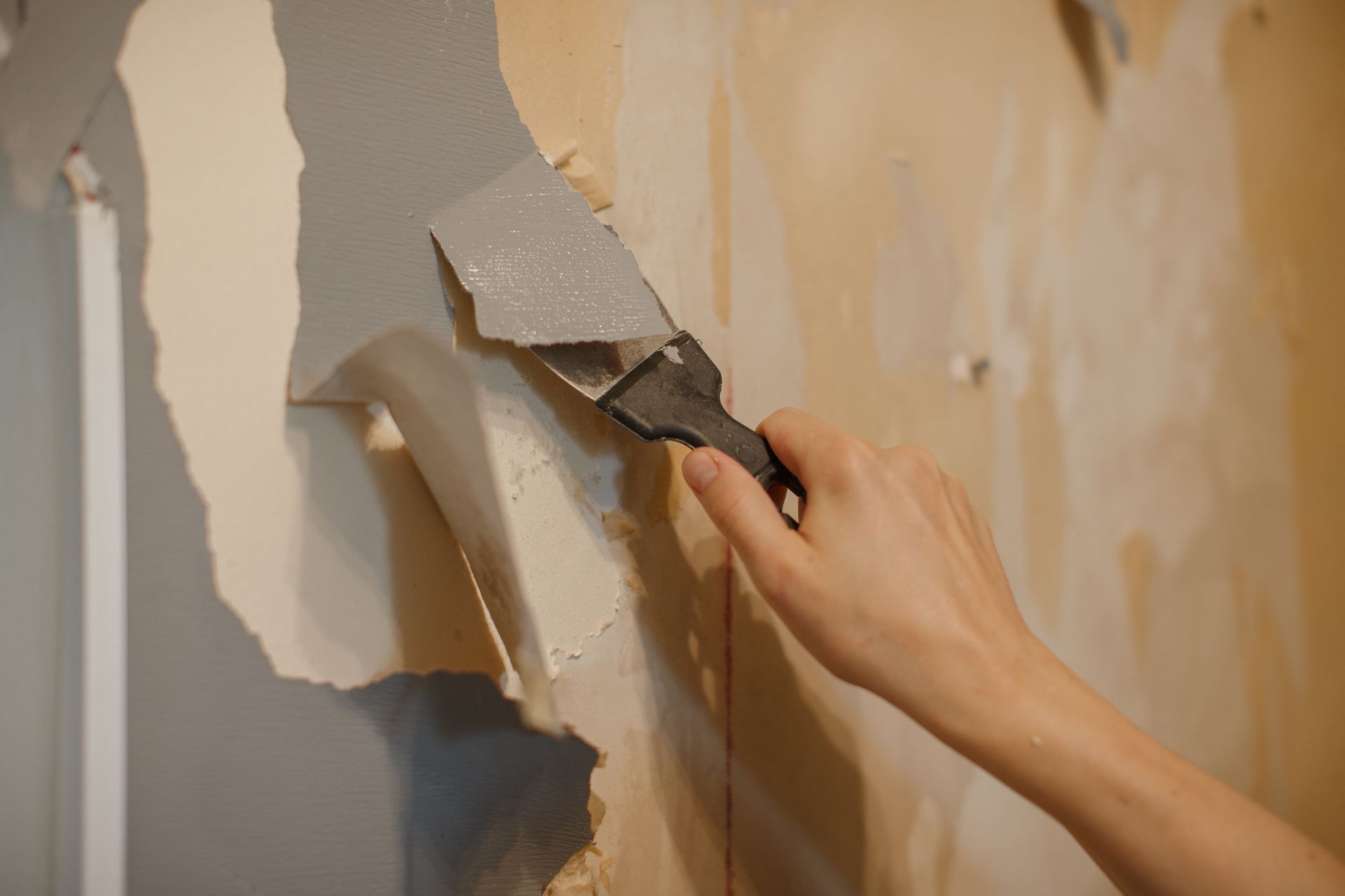 Hand using a putty knife to remove peeling wallpaper from a wall.